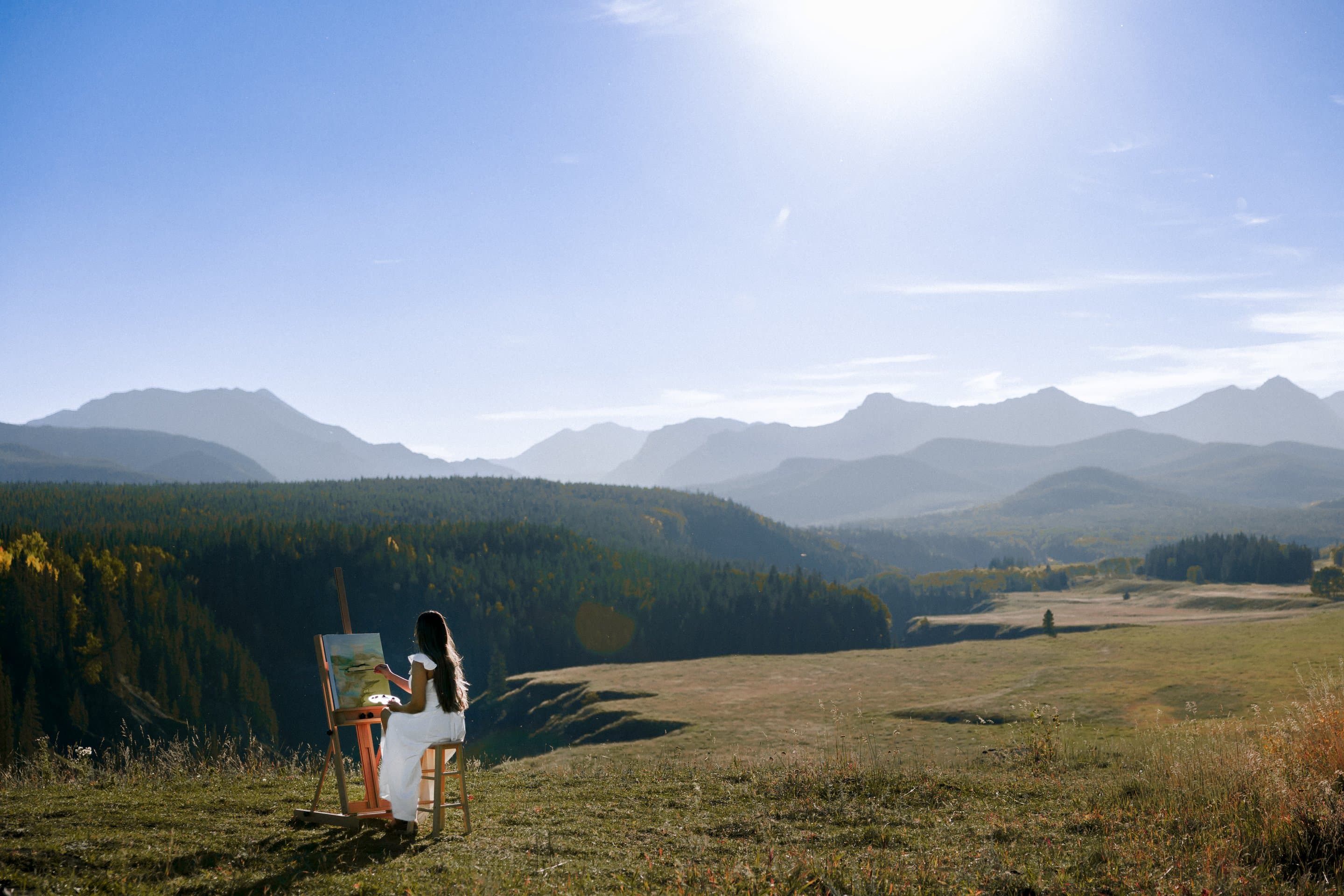 Kiran Rai-Bhullar painting at an easel on a mountain meadow, forest and ranges behind her. Photograph by Galyna Gaponiuk.