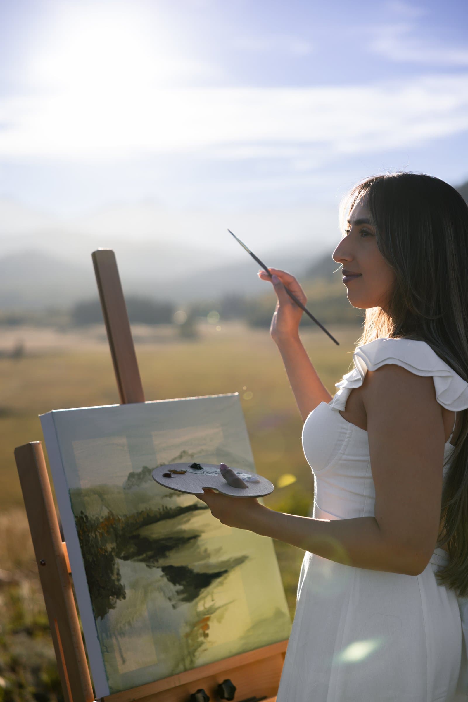 Kiran Rai-Bhullar painting at her easel in a mountain meadow, lit from behind by the evening sun. Photograph by Galyna Gaponiuk.