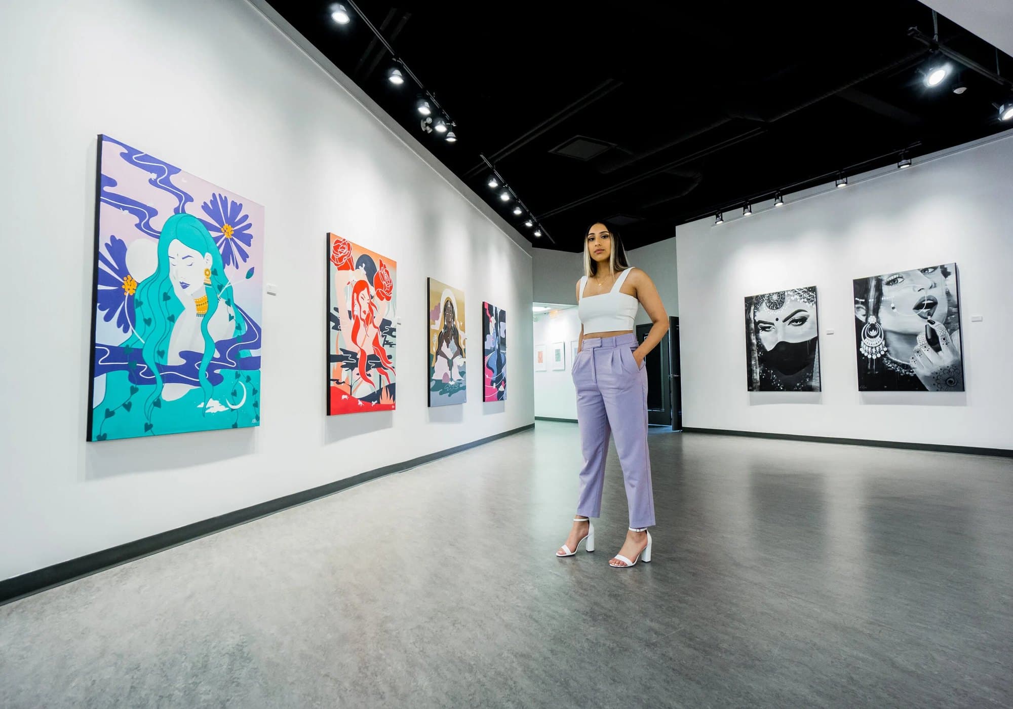 Kiran Rai-Bhullar standing in The Hub Gallery at artsPlace, the four Maharani Mentality paintings on the wall to the left and two black and white portraits to the right