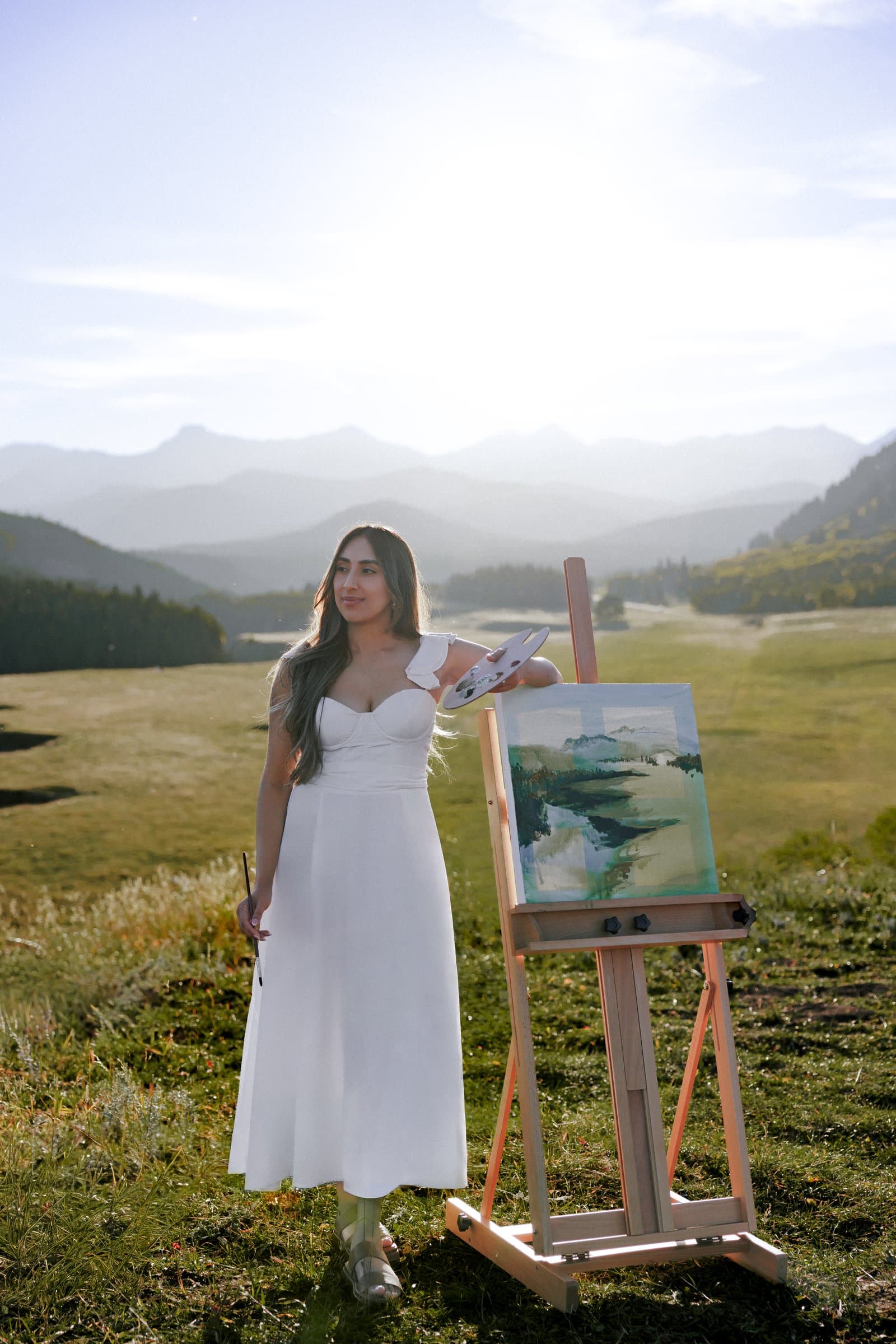 Kiran Rai-Bhullar in a white dress at her easel in a mountain meadow, palette in hand