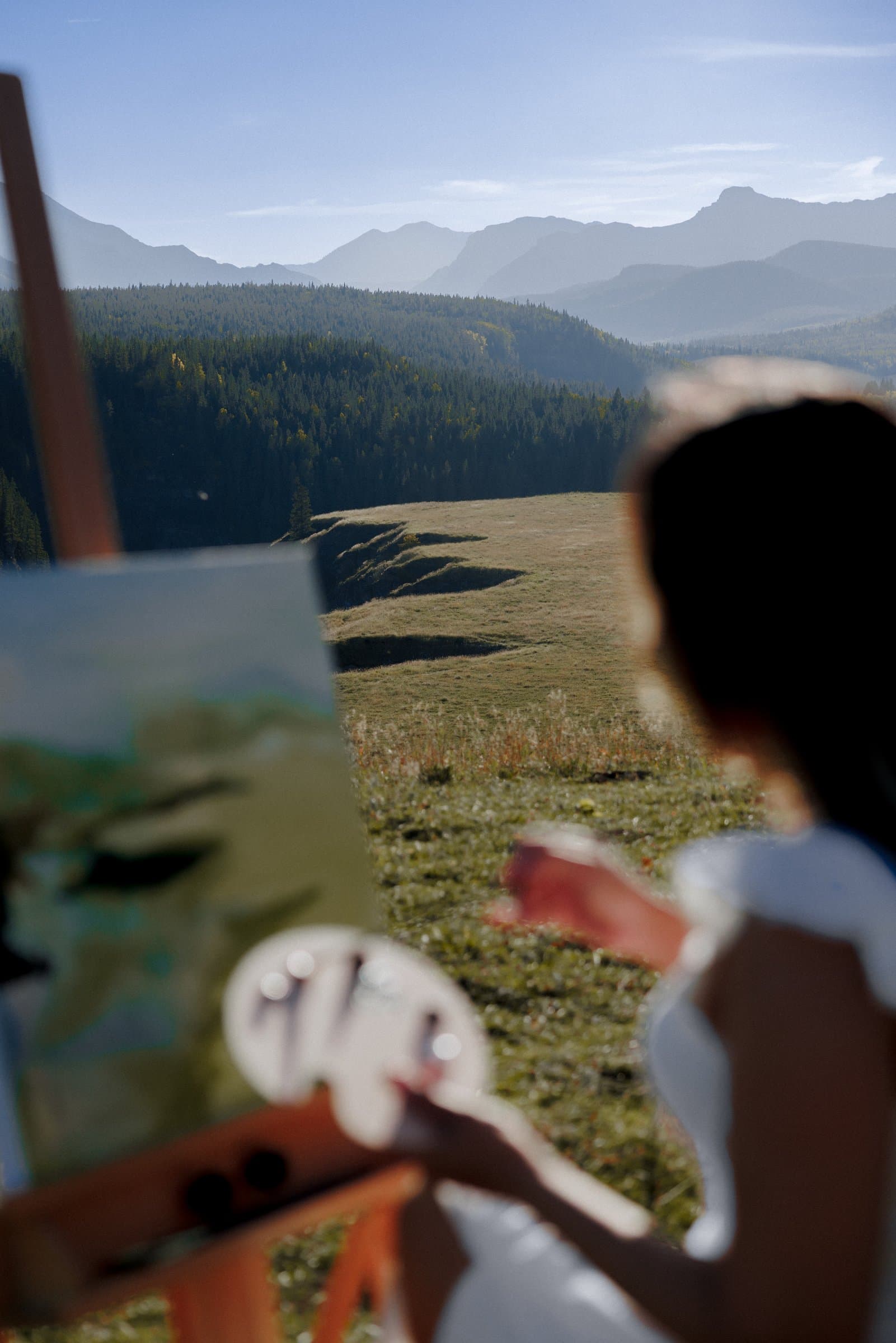 Kiran Rai-Bhullar at her easel in a mountain meadow, seen softly from behind, with forest and ranges in the distance. Photograph by Galyna Gaponiuk.