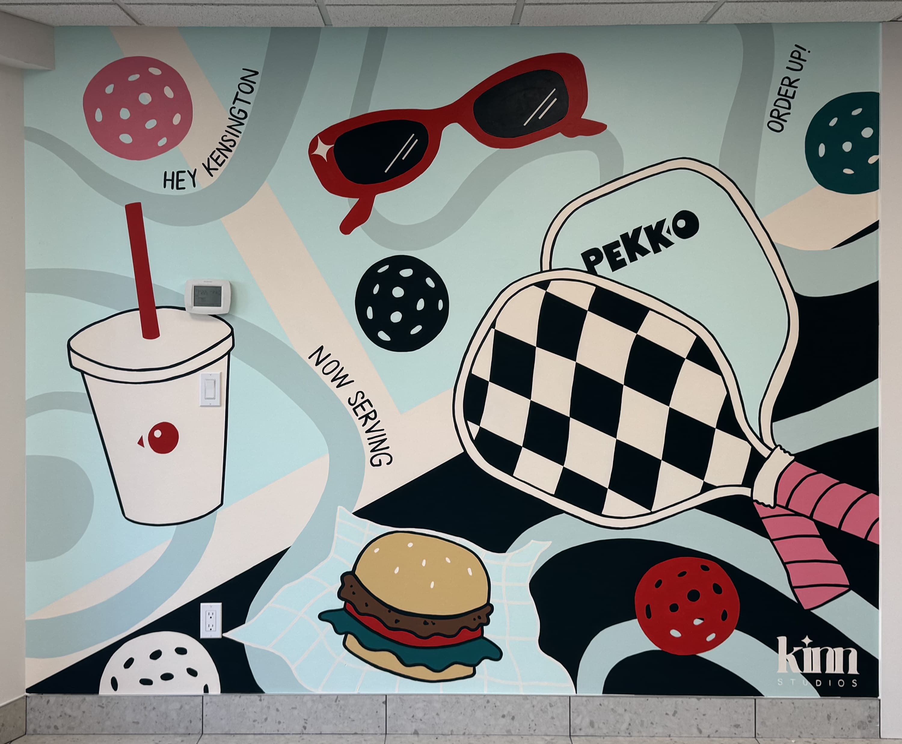 Pekko's other dining-room wall: a checkerboard pickleball paddle with the PEKKO logo, pickleballs, red sunglasses, a drink and a burger, with HEY KENSINGTON, NOW SERVING and ORDER UP! lettering